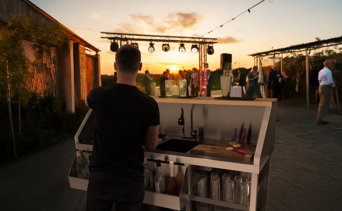 En bartender i svart t-shirt står vid en modern bar med glas och flaskor, med en solnedgång och festlig atmosfär i bakgrunden