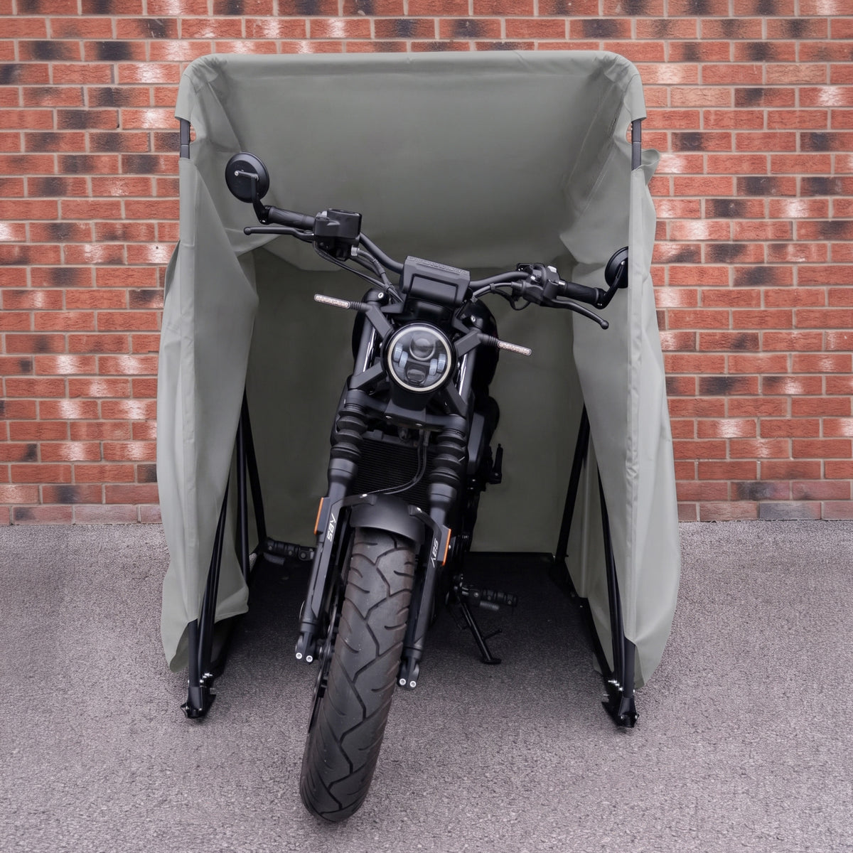 Svart motorcykel skyddad av en grön presenning, placerad framför en tegelvägg, med fokus på detaljer och robust design