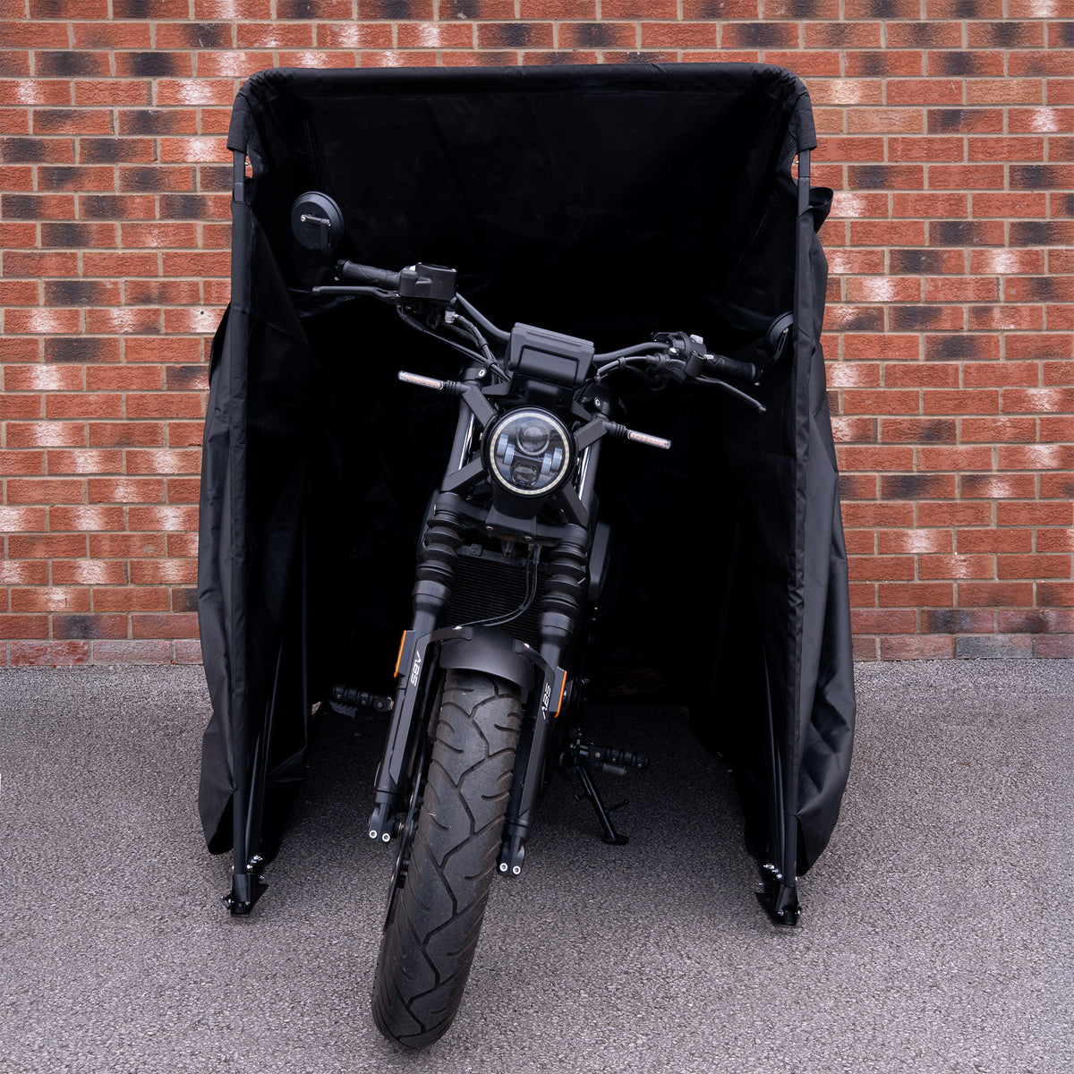 Svart motorcykel med kraftig däckprofil står skyddad i en svart täckning mot en tegelvägg i bakgrunden