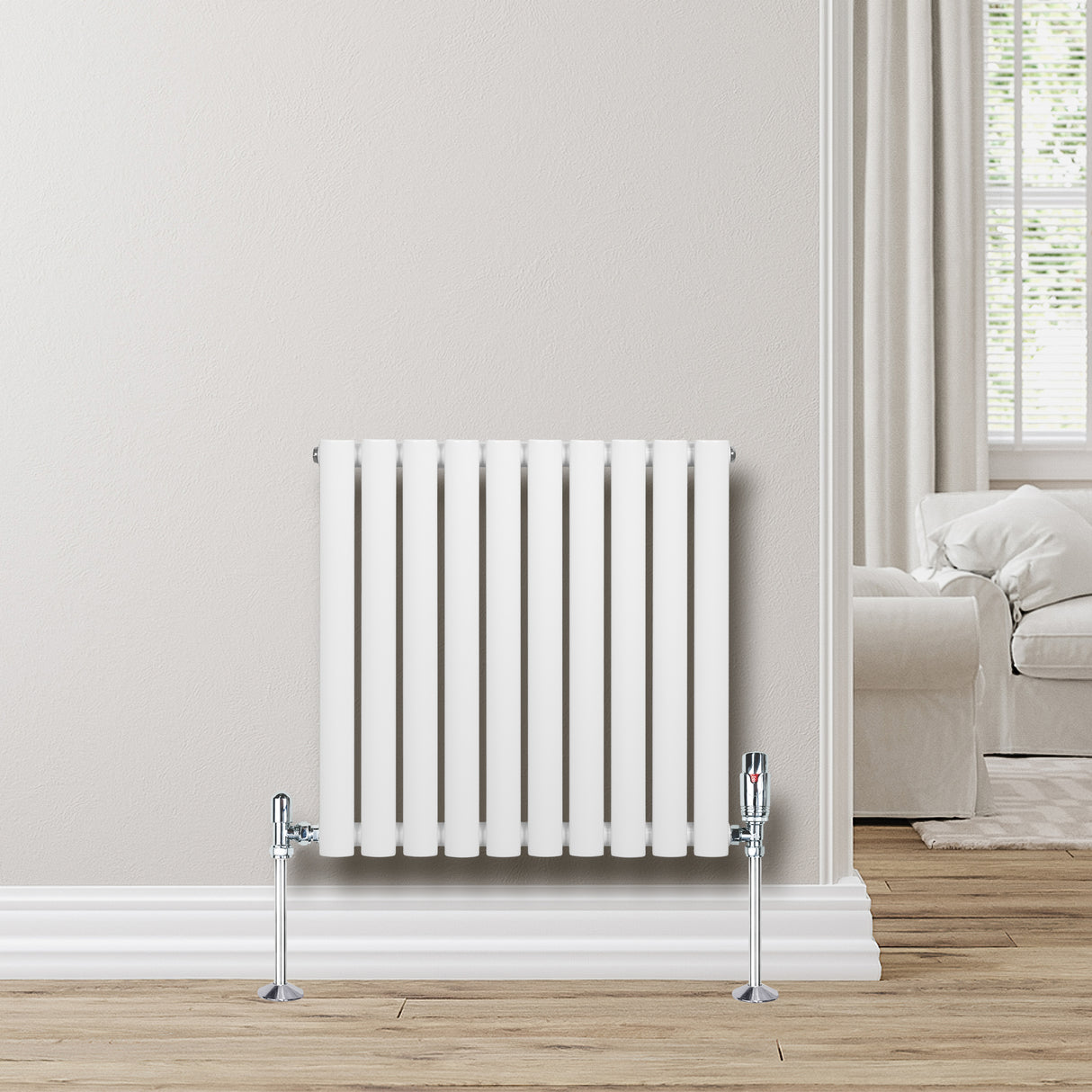 Oval vit enkelpelarradiator med kromade ventiler, placerad på en ljus vägg med trälookgolv och mjuka gardiner i bakgrunden