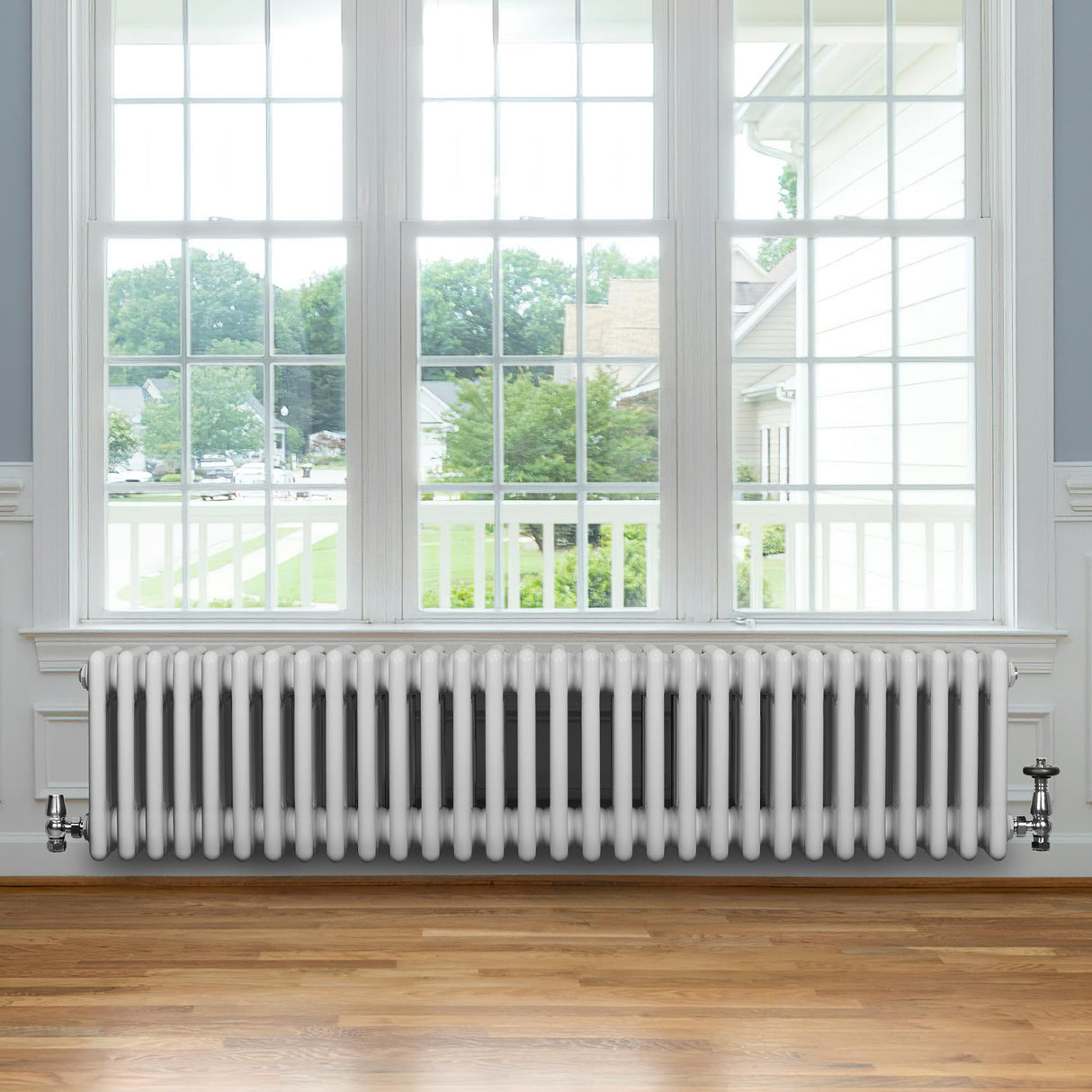 Traditionell vit 4-kolonnsradiator i metall, placerad under ett stort fönster med trädetaljer och synlig utsikt utomhus