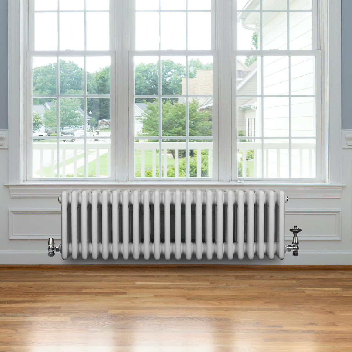 Traditionell vit 4-kolonnsradiator i metall, placerad under ett stort fönster med trädetaljer och ljus träparkettgolv