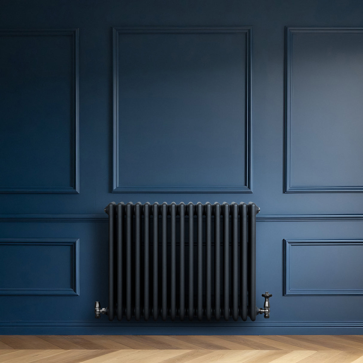 Traditionell antracitgrå 4-kolonnsradiator med släta ytor och runda kanter, installerad på en mörkblå vägg med träparkettgolv