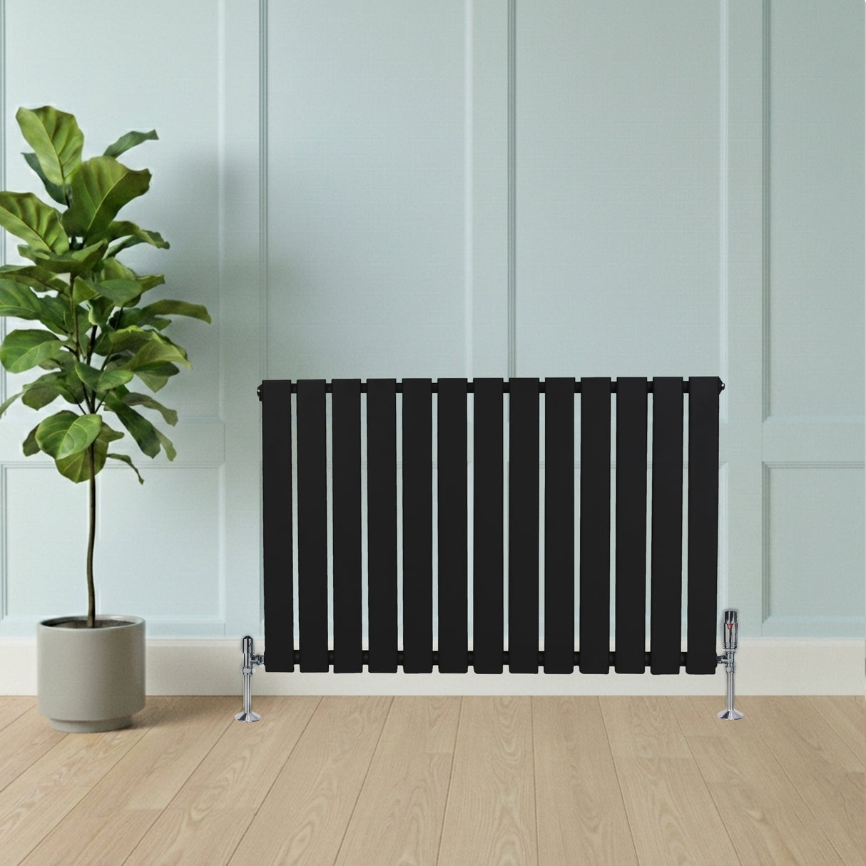 Svart platt enkelpelarradiator med vertikala ribbor, placerad framför en grön växt i en ljus och modern inredning