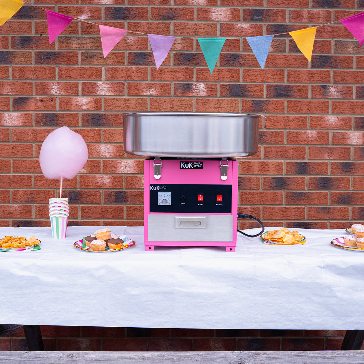 KuKoo sockervaddsmaskin i rosa och silver, placerad på ett bord med festlig dekoration, omgiven av godis och cupcakes