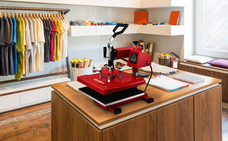 En röd värmepress står på ett träbord i en kreativ butik, omgiven av färgglada t-shirts och material för DIY-projekt