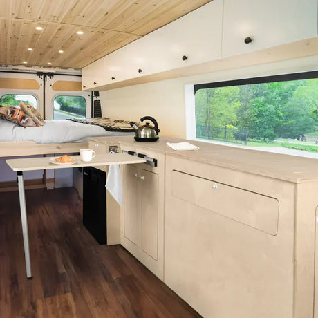 Ljust inrett campervan med träpanel, köksdel, säng och fönster som ger utsikt över grönskande landskap och naturligt ljus