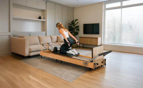 Kvinna i sportkläder utför Reformer Pilates på en träbänk i ett ljust rum med en soffa och stor fönster för naturligt ljus