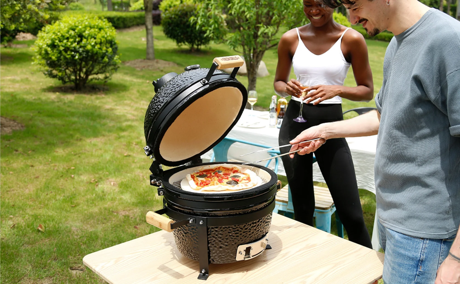 En svart kamado-grill med en öppen lock, som grillas pizza med färgglada pålägg, omgiven av grönska och glada människor
