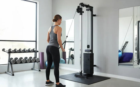 Kvinna i grå träningstopp använder väggmonterad kabelmaskin i ett ljust gym med vikter och spegel i bakgrunden