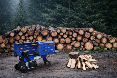 En blå vedklyv står på en grusig yta framför en hög av staplade vedträn och en hög med klyvna vedbitar i skogen