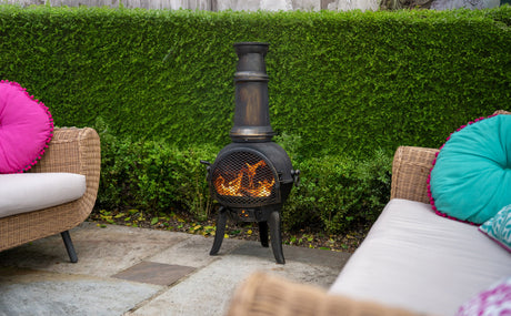 Svart chiminea med eld som brinner, omgiven av grön häck och bekväma soffor med färgglada kuddar i rosa och turkos