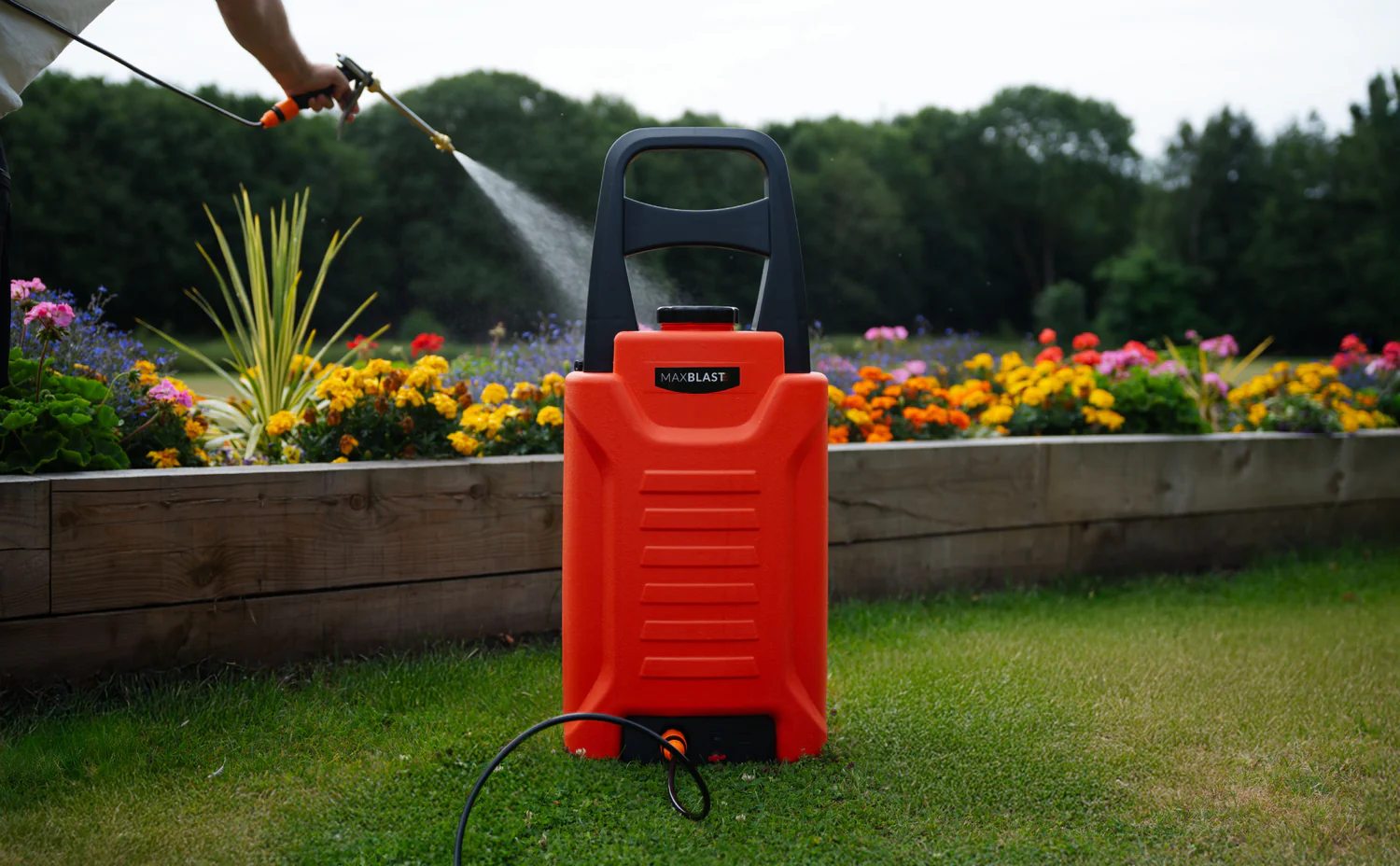 Röd trädgårdsspruta står på gräset framför en färgglad blomrabatt med blommor i olika nyanser och en person som sprayar