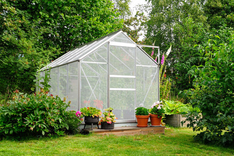Ett växthus med transparenta väggar, omgiven av gröna växter och färgglada blommor i krukor, på en grön gräsmatta