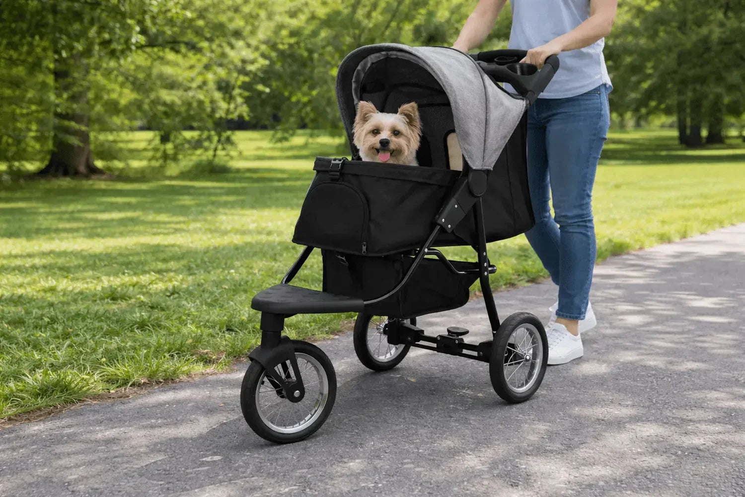 En person går med en svart joggingvagn för husdjur, där en glad hund sitter bekvämt inuti, omgiven av grönska och träd
