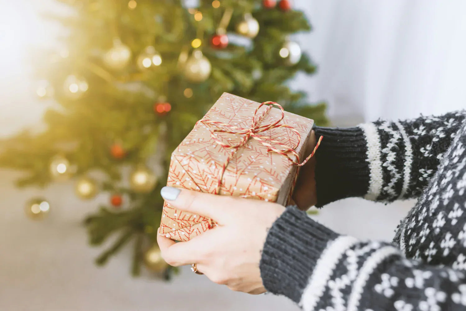 En person håller ett vackert inslaget julklapp i guldigt papper med röd snöre framför en julgran med glittrande dekorationer