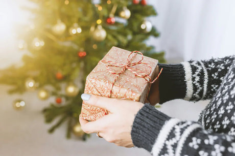 En person håller ett vackert inslaget julklapp i guldigt papper med röd snöre framför en julgran med glittrande dekorationer