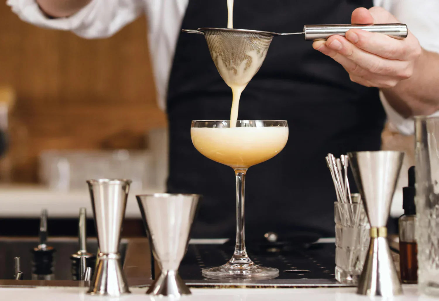 En bartender häller en krämig cocktail genom en sil i ett elegant cocktailglas, omgiven av mätverktyg och barutrustning