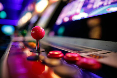 Rosa joystick och knappar på en retro arkadmaskin, med suddig bakgrund av färgglada skärmar och mjukt ljus