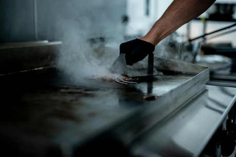 En hand i svart handske vänder kött på en glänsande elektrisk stekhäll, omgiven av ånga och en varm, metallisk yta
