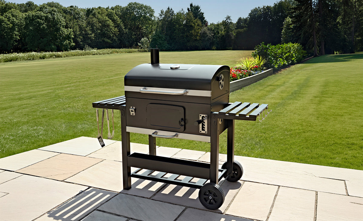 Svart kolgrill med två sidobord, placerad på en stenbelagd uteplats med grön gräsmatta och träd i bakgrunden