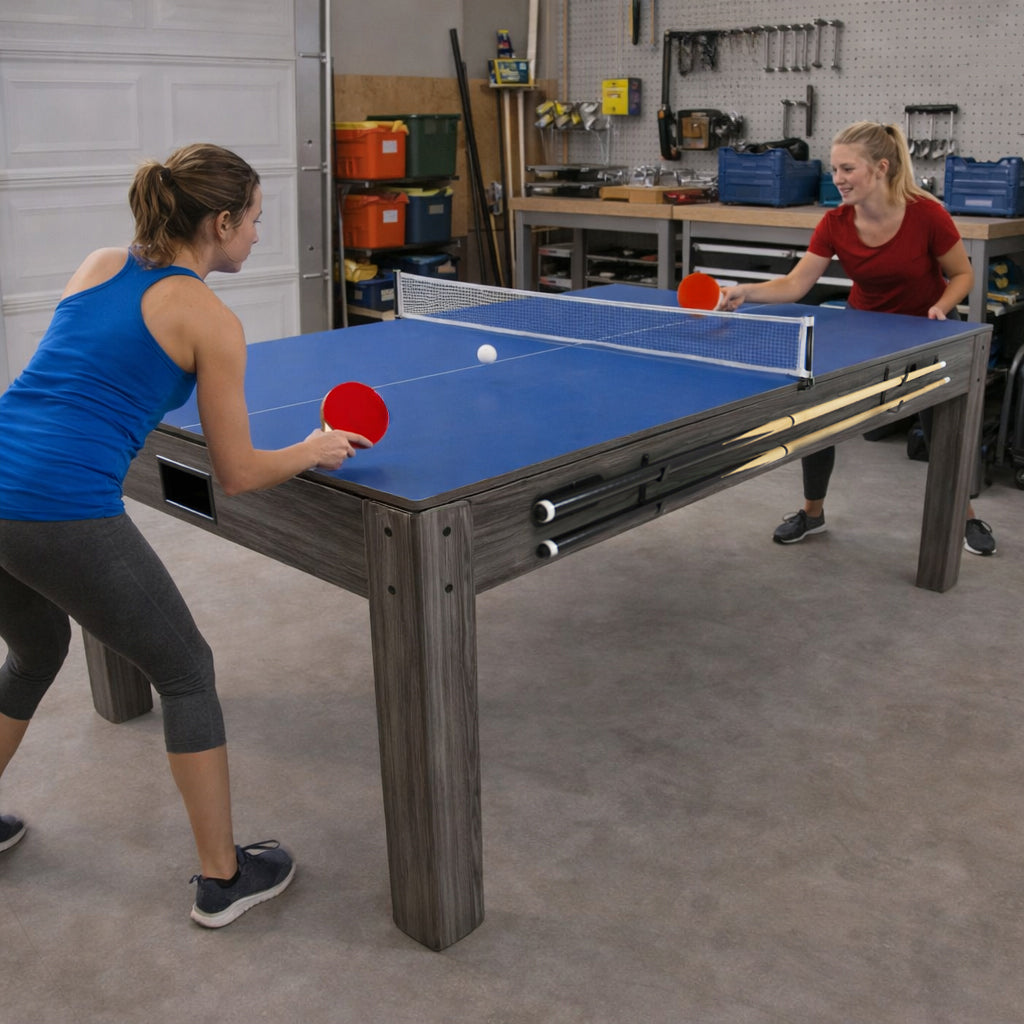 Spelbord i grått med blått bord och förvaringsbänk, två personer spelar bordtennis med racketar och boll i garage-miljö