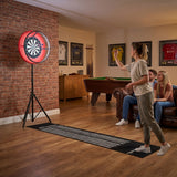 Grått spelbord med dartpaket och LED-lampa, en kvinna kastar dartpilar mot en röd darttavla i en mysig inomhusmiljö