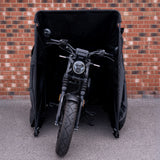 Svart motorcykel med kraftig däckprofil står skyddad i en svart täckning mot en tegelvägg i bakgrunden