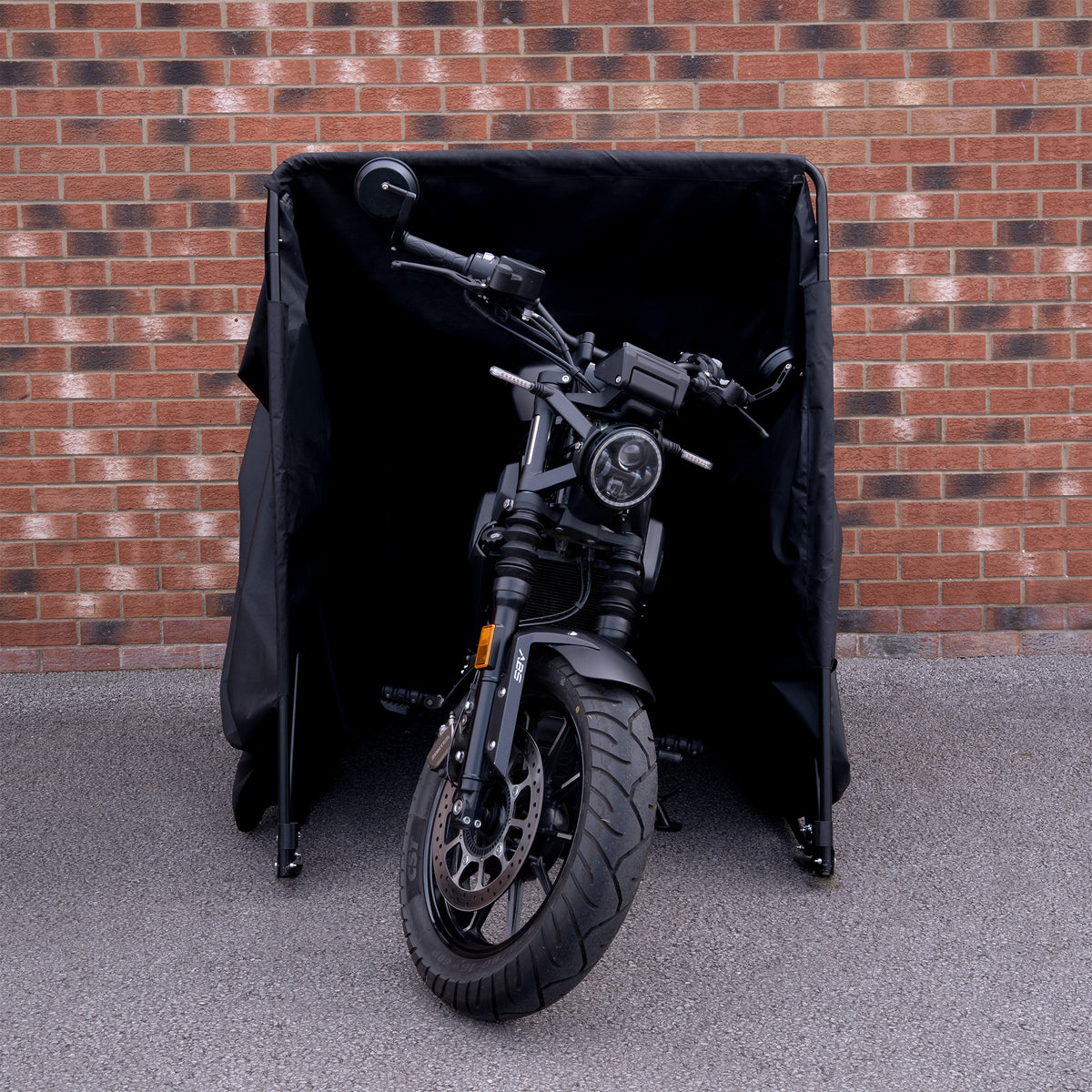 Svart motorcykel delvis skyddad av en svart täckställning, med en tegelvägg i bakgrunden och en grå asfalt under