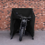 Svart motorcykel skyddad av ett väderskydd, stående framför en tegelvägg, med synlig däckmönster och detaljerad design