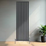 Platt enkelkolumnsradiator i antracitgrå med vertikala ribbor, placerad på en ljus träyta med en grön växt i bakgrunden