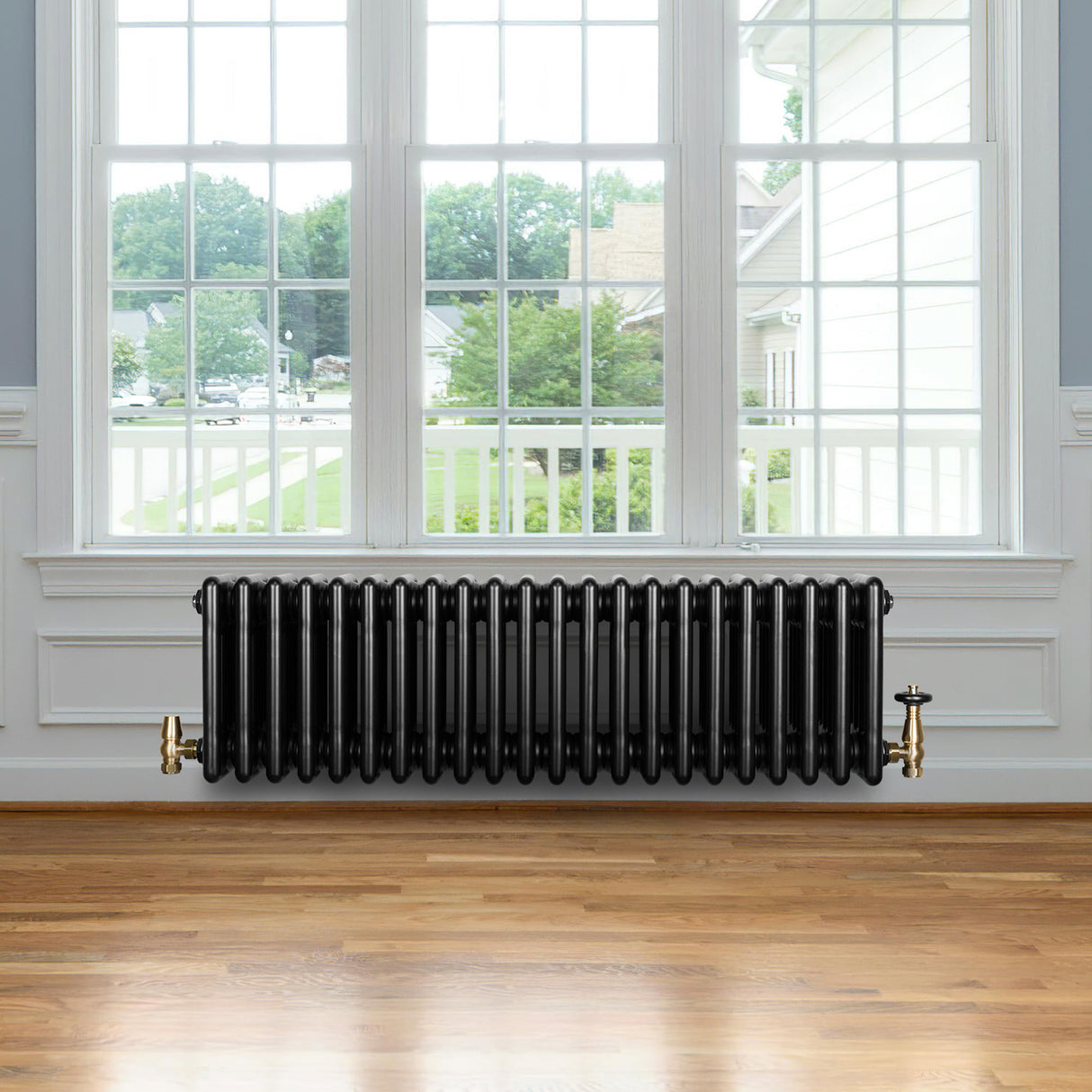 Svart traditionell 4-kolonnsradiator i metall med ventiler, placerad under ett stort fönster i ett ljust rum med trägolv