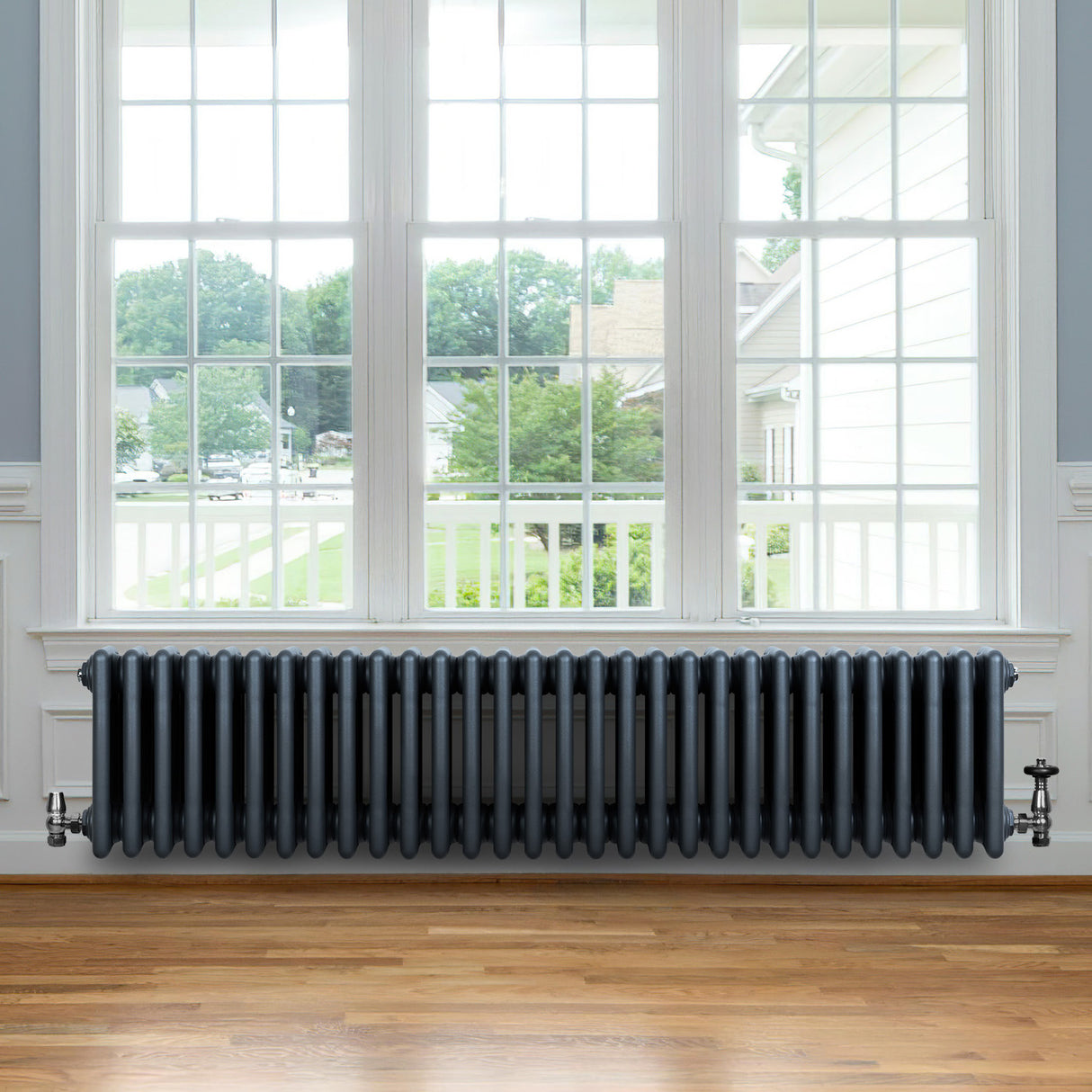 Traditionell antracitgrå 4-kolonnsradiator med släta ytor, placerad under ett stort fönster med trädetaljer och parkettgolv