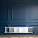 Traditionell vit 4-kolonnsradiator i metall, installerad mot en mörkblå vägg, med eleganta, rundade former och synliga ventiler