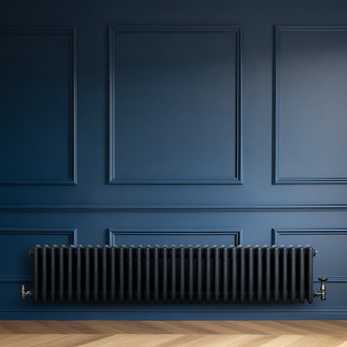 Traditionell antracitgrå 4-kolonnsradiator i metall med släta ytor, placerad mot en mörkblå vägg och ett ljust trägolv