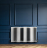 Traditionell vit 4-kolonnsradiator i metall, placerad mot en mörkblå vägg, med släta, runda rör och eleganta ventiler