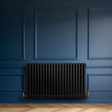 Svart 4-kolonnsradiator med glänsande yta och ventiler, placerad mot en mörkblå vägg med eleganta paneler och träplankor på golvet