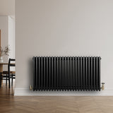 Svart traditionell 4-kolonnsradiator med glänsande yta, installerad på en ljus vägg i ett modernt rum med trädetaljer