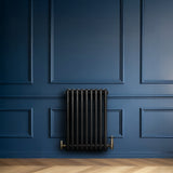 Svart traditionell 4-kolonnsradiator i metall, placerad mot en mörkblå vägg med eleganta paneler och ett ljust trägolv