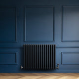 Traditionell antracitgrå 4-kolonnsradiator med släta ytor och runda kanter, installerad på en mörkblå vägg med träparkettgolv