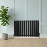Svart platt enkelpelarradiator med vertikala ribbor, placerad framför en grön växt i en ljus och modern inredning