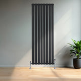 Svart platt enkelpelarradiator i metall med vertikala ribbor, placerad mot en ljus vägg och omgiven av en grön växt i kruka
