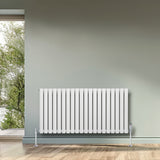 Oval enkelpelarradiator i vit färg, 600 x 1200 mm, med släta ytor och stående ribbor, placerad mot en grön vägg nära ett fönster