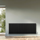 Svart oval enkelpelarradiator med släta ytor, placerad mot en ljus vägg, med träparkett och fönster som släpper in naturligt ljus