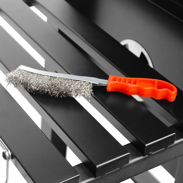 Rostfri stålborste med orange handtag, placerad på en svart metallhylla, perfekt för rengöring av grill och köksutrustning