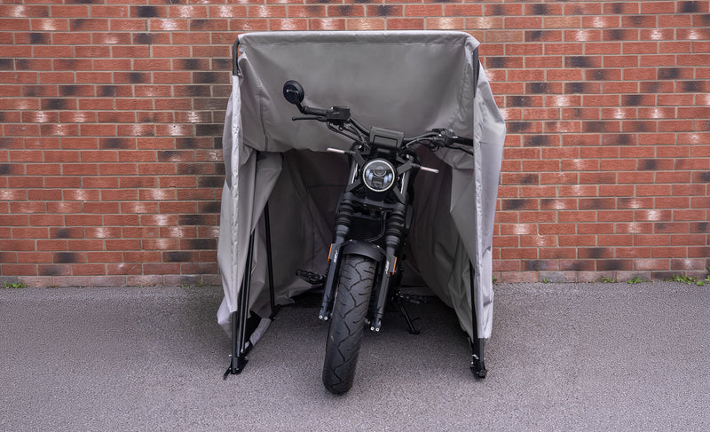 Svart motorcykel skyddad av en grå presenning, placerad framför en tegelvägg, med fokus på däck och styre