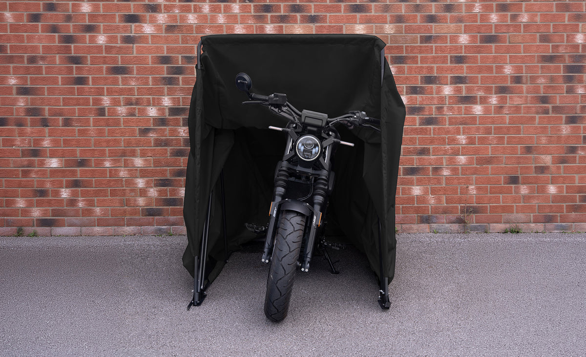 En svart motorcykel skyddad av ett väderskydd, placerad framför en tegelvägg, med synlig däck och styre