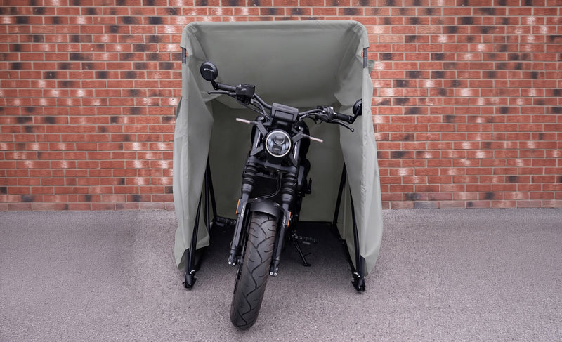 Svart motorcykel med kraftig däckprofil står skyddad under en grön, vattentät täckställning mot en tegelvägg