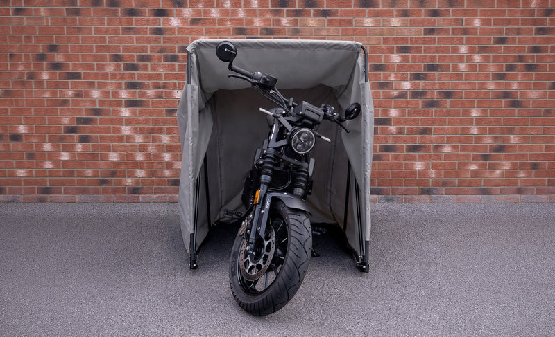 En grå motorcykel står i ett skyddande tält med en tegelvägg i bakgrunden, däckets mönster och detaljer syns tydligt