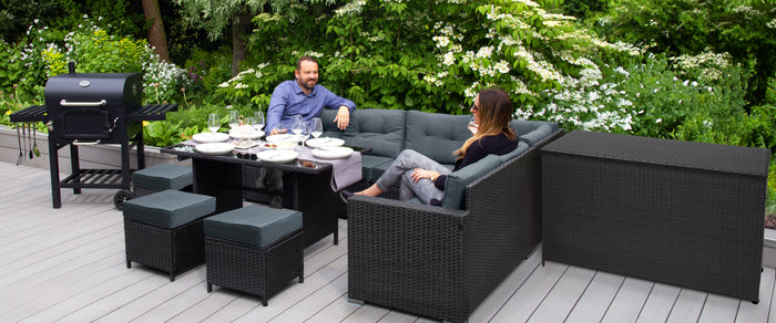 Elegant utemöbelset i svart rotting med grå kuddar, en matplats med tallrikar och en grill i bakgrunden, omgiven av grönska